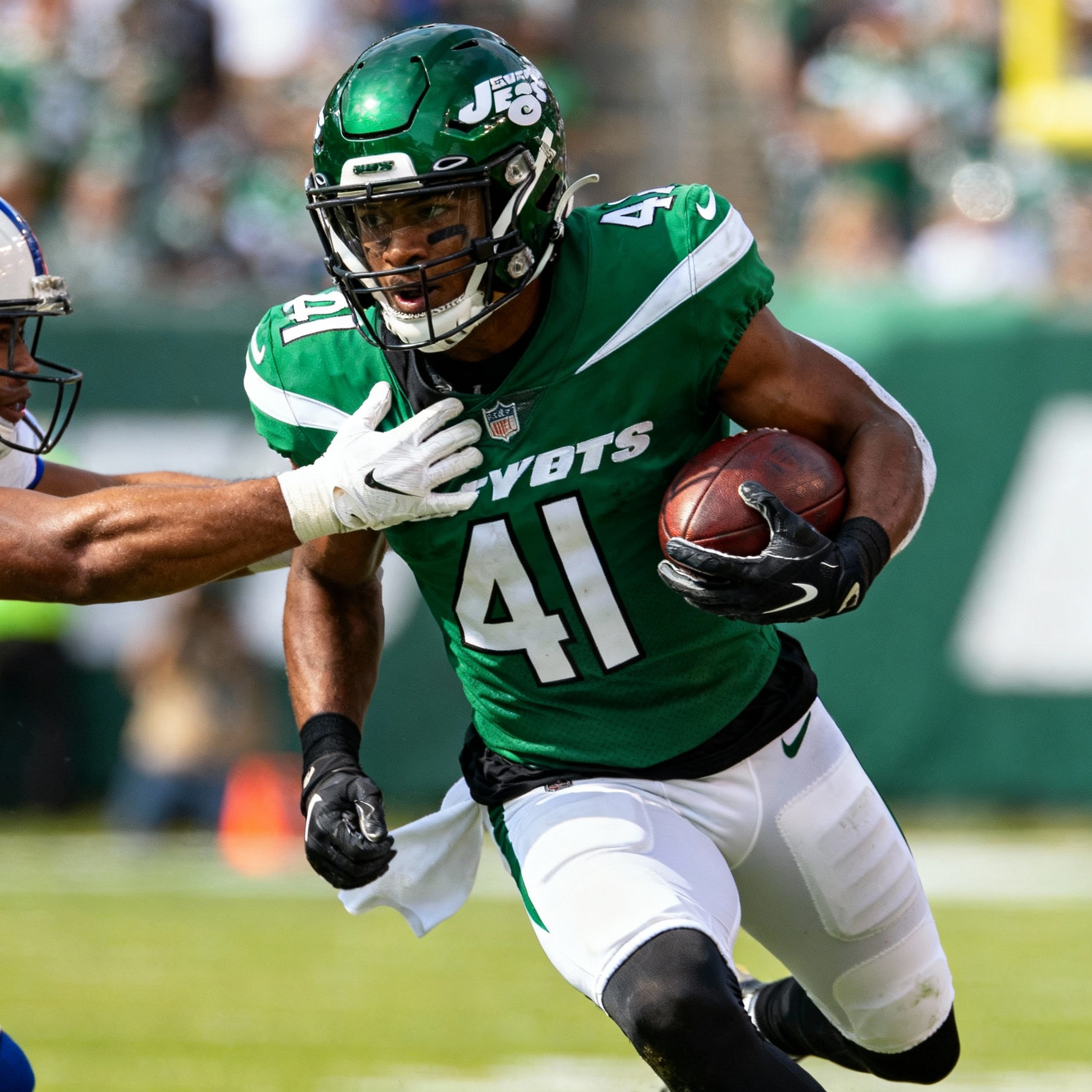 “Breece Hall in a green New York Jets uniform evading a tackle while sprinting forward with the football in his right arm on the field.”