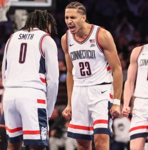 Dynamic action shot of UConn Huskies men's basketball player in white and navy uniform driving to the basket at Madison Square Garden during the 2025 Jimmy V Classic, with blurred crowd and teammates symbolizing team depth and national title contention.